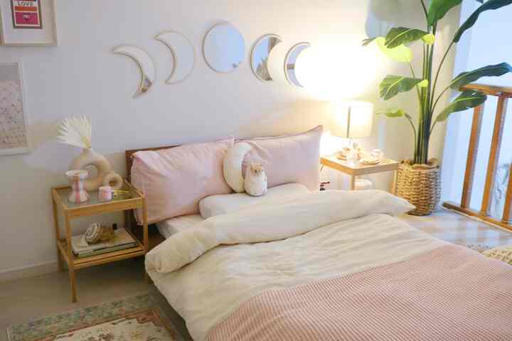 パステルピンクとホワイトの色調の寝室、木製ナイトテーブルと大きな植物が配置された柔らかく自然な雰囲気のインテリア