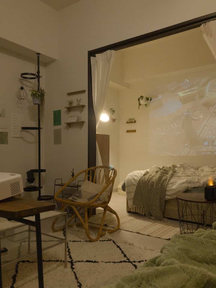 ベージュ色調の1LDKスタジオ、モダンなラタンチェアとプロジェクターが配置された寝室兼リビングの雰囲気のあるインテリア