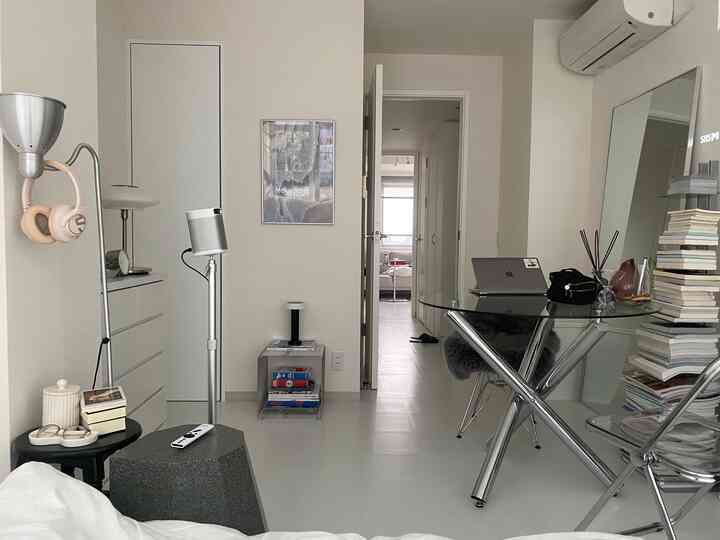 Bright white-walled bedroom with glass table hosting a laptop and stool, showcasing a minimalist and modern home office setup