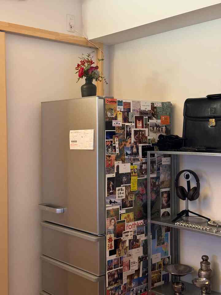 A kitchen space featuring a silver refrigerator and metal shelving unit with flowers on top and various pictures stuck on the wall beside it
