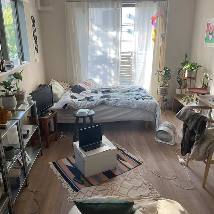 ホワイトとウッド調が調和するコンパクトな寝室。シンプルなベッドとデスク、多数の植物や収納アイテムが配置された雰囲気のあるインテリア