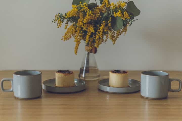 ナチュラルカラーのダイニングテーブルに陶器のマグカップと皿、黄色い花の花瓶が配置されたカフェ風の印象的な空間