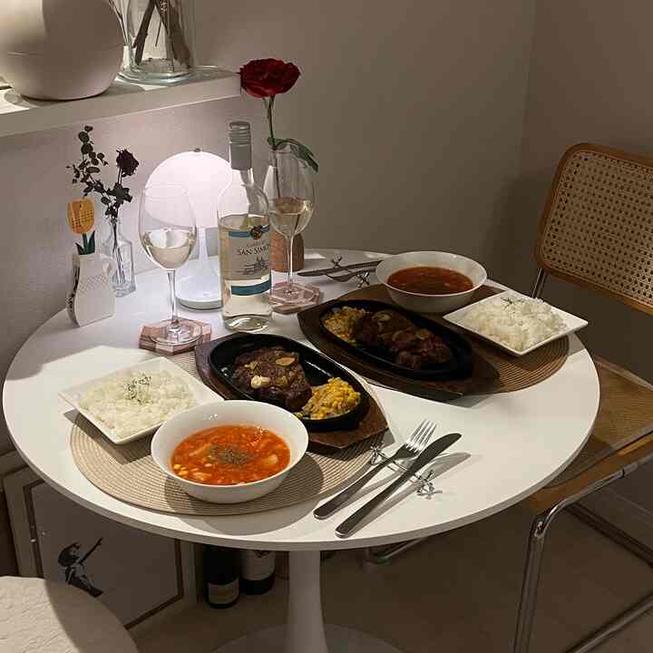 Cozy natural Nordic dining space featuring a white round dining table and rattan chair, set with steak and wine for dinner