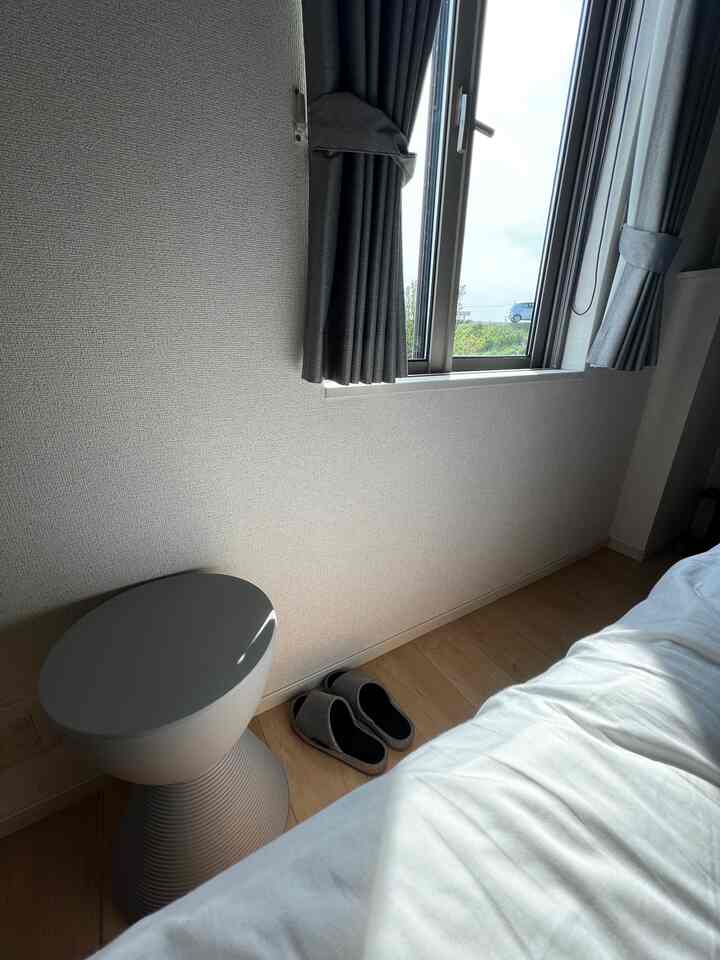 Bright bedroom with white bedding, gray curtains, and a stool near the window with natural sunlight