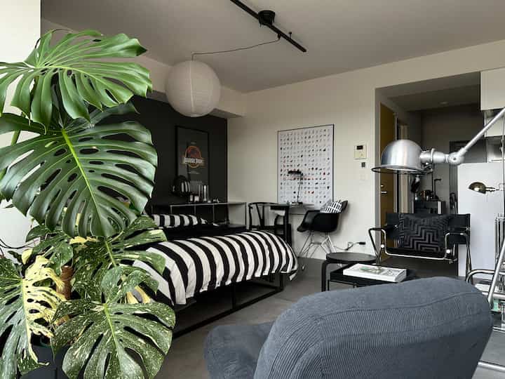 グレーと黒を基調としたワンルームの寝室、コンパクトな空間にベッド、デスク、アームチェアが配置されたモダンでミニマルなインテリア