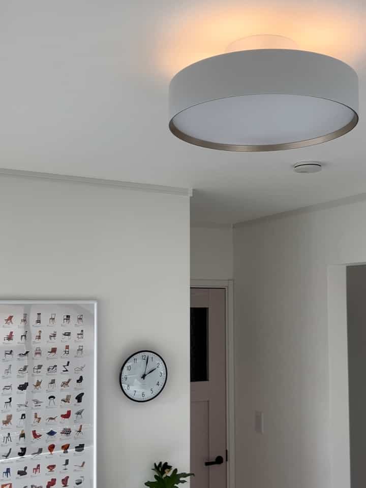 白い壁と扉、間接照明付きの大きなシーリングライト、壁掛けの黒い時計があるシンプルな玄関空間