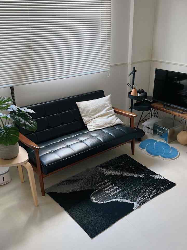 白い壁と床に、黒のレザー製2人掛けソファとナチュラルな木製サイドテーブル、植物が配置された温かみのあるリビング空間