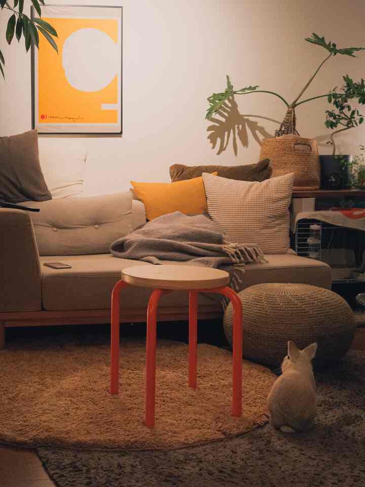 ナチュラルな色調の1LDKリビング、ベージュのソファとオレンジの脚のスツール、クッションや植物が配された雰囲気のある空間
