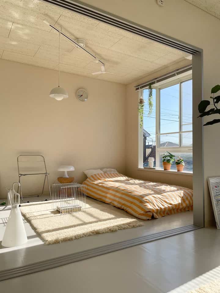 クリーム色調の小さな寝室、窓辺に置かれた低いベッドと植物が配置された雰囲気のあるインテリア