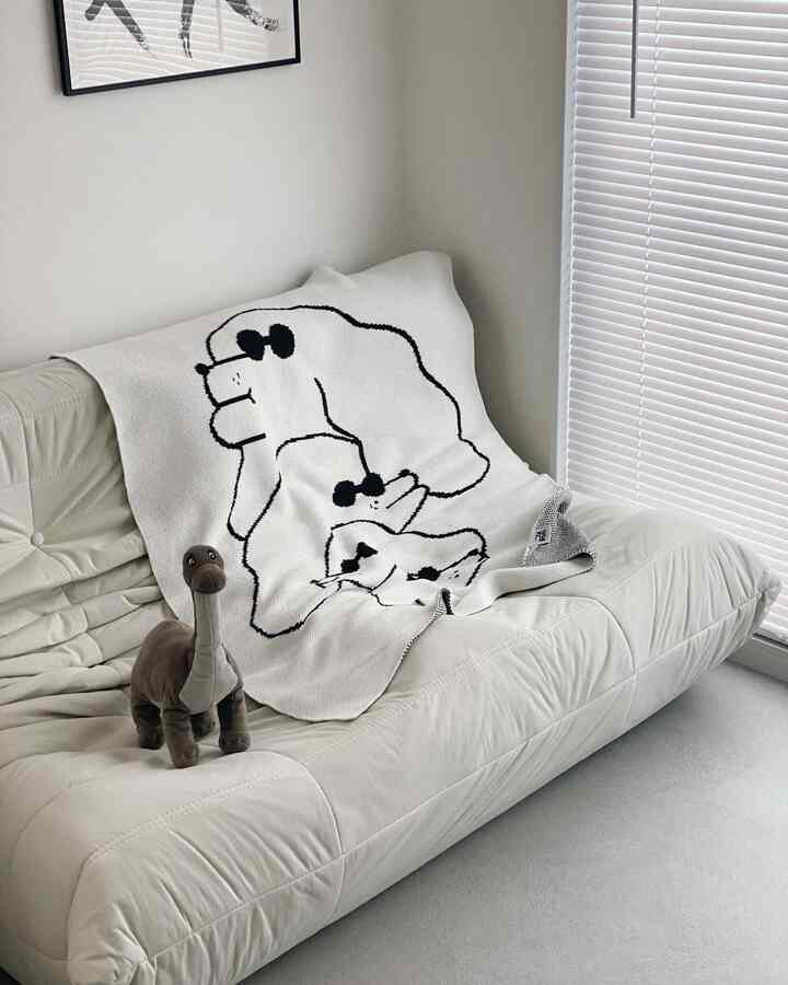 White-toned living room featuring a cream sofa with a dinosaur-patterned blanket and plush toy on a simple, minimalist setting