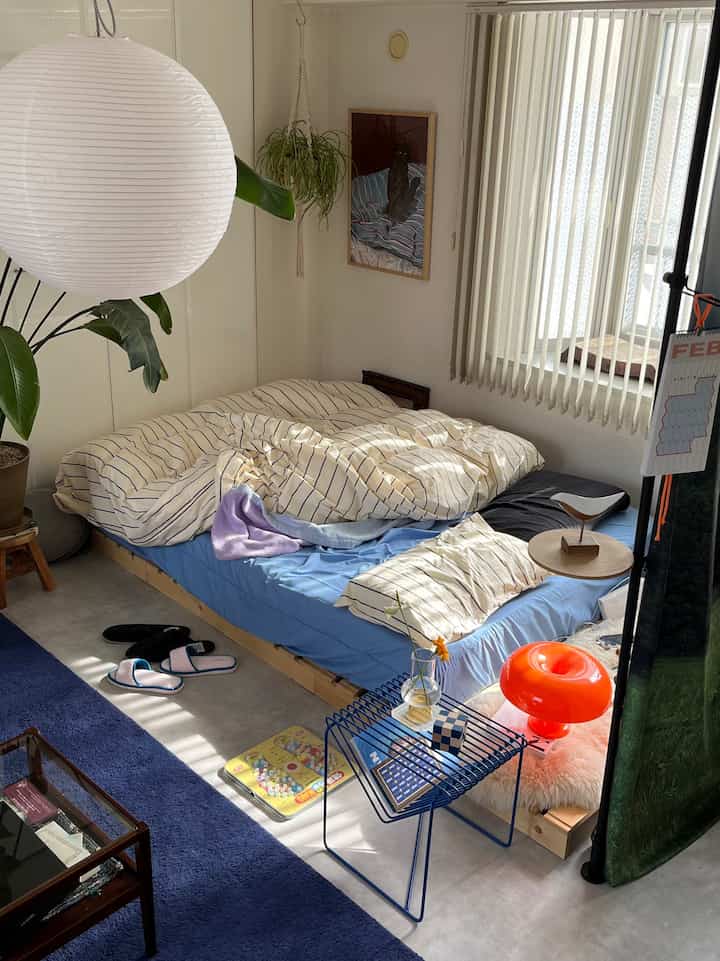 ホワイトとブルーの色調のコンパクトな寝室、木製ベッドとブルーのサイドテーブル、ホワイトのフロアランプと植物が配置された自然な雰囲気のあるインテリア