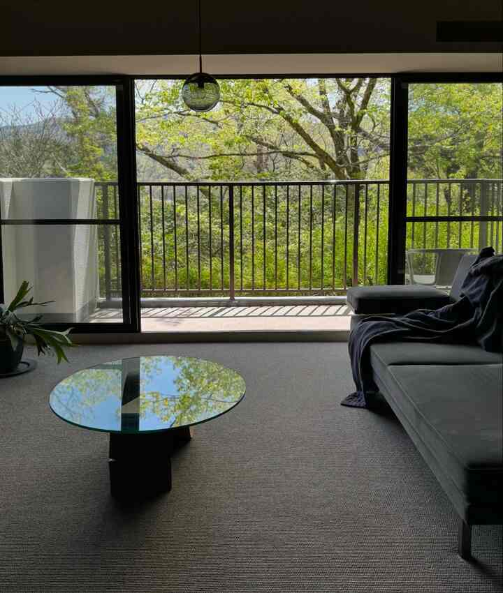 A natural-toned living room with a round glass coffee table on grey carpet, dark gray sofa on the right, and green trees visible beyond the balcony