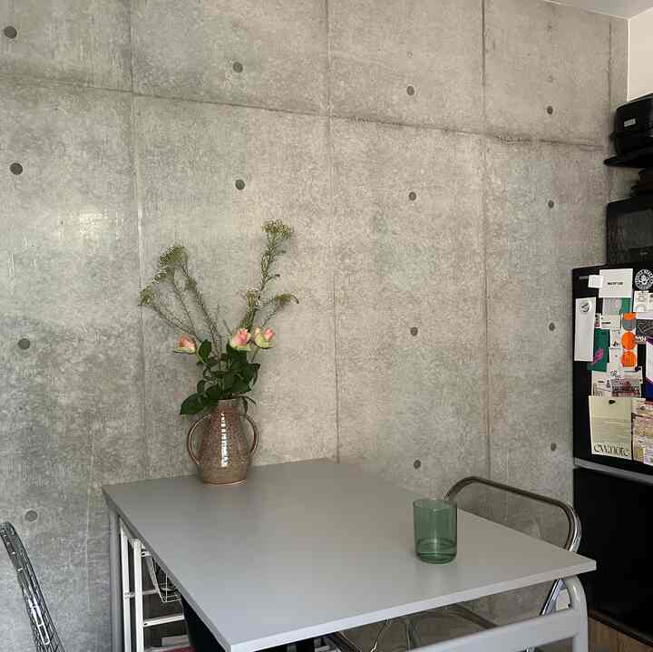 グレーのコンクリート壁と透明なダイニングチェアがあるコンパクトなキッチンスペース、テーブルの上に花瓶が配置されたモダンな雰囲気