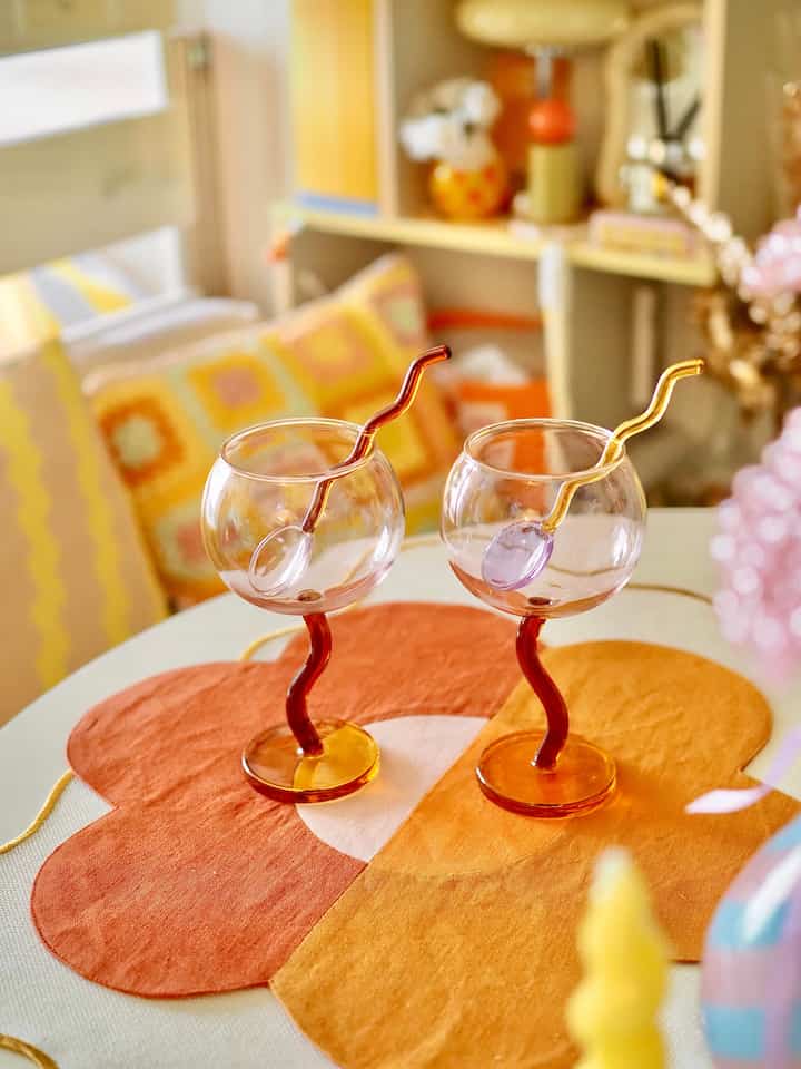 暖かい黄色とオレンジのトーンのダイニングテーブルに、うねった脚とスプーンが付いた透明なゴブレットが2つ並ぶ、雰囲気のあるカフェ風インテリア