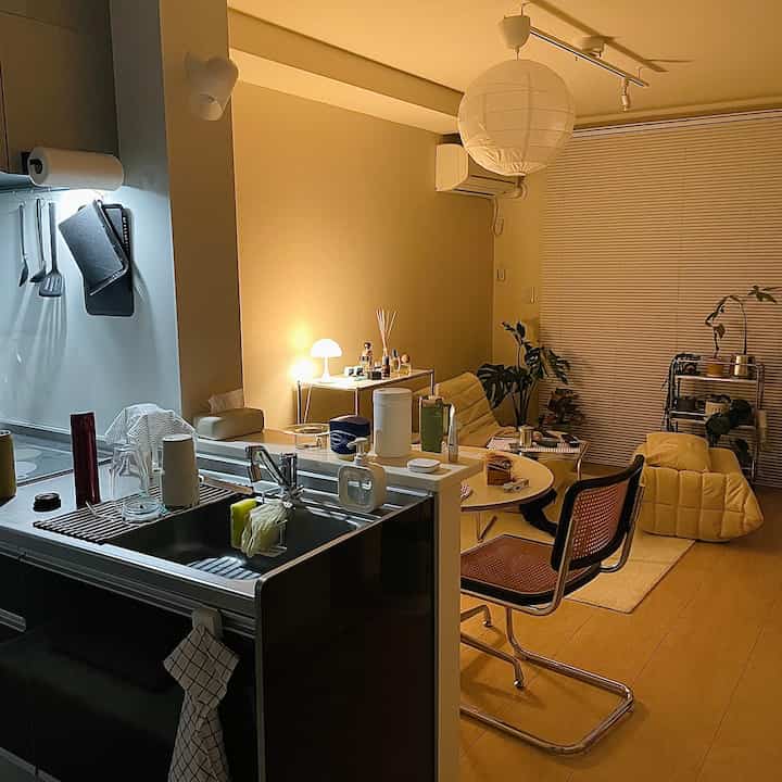 Warm yellow-toned single household apartment living room and kitchen featuring a rattan chair, round dining table, diffuser, and table lamp in a neat cozy setting