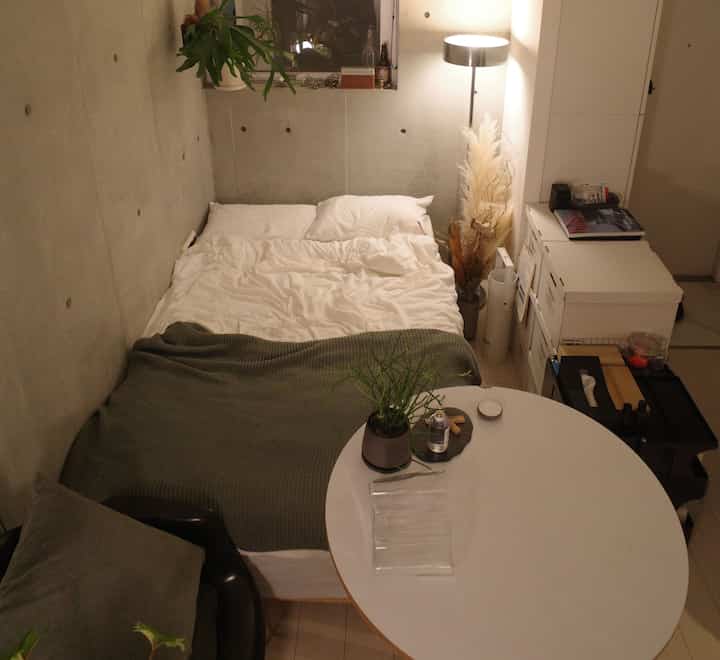 ホワイトとグリーンのトーンが映える6畳の寝室。快適なベッドと丸テーブル、フロアランプ、自然な装飾が配置された居心地の良いワンルーム