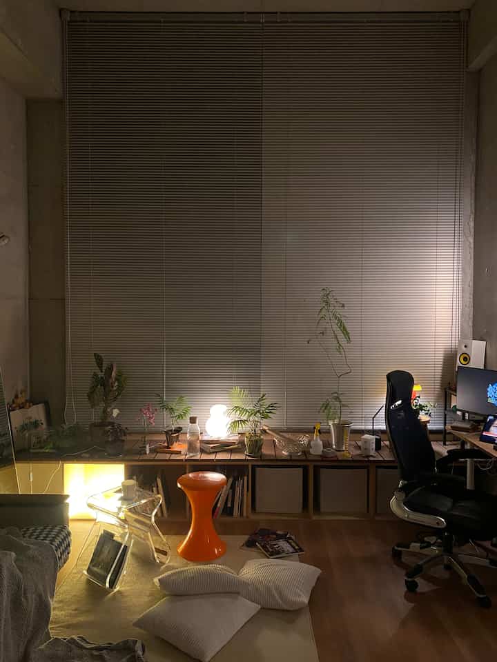 ナチュラルカラーのリビング兼ホームオフィス、オレンジのスツールとオフィスチェア、多数の植物が配置された落ち着いた空間