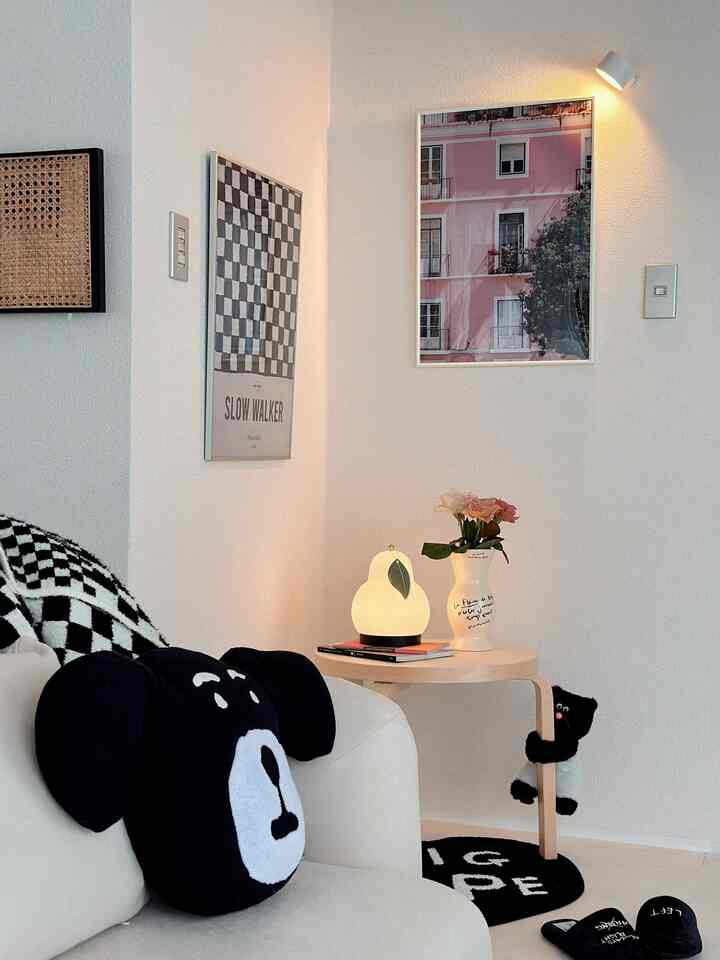 明るいベージュのソファと丸い木製スツールの上に置かれた梨型のテーブルランプ、白い壁に2つのポスターがあるシンプルでナチュラルなリビング空間