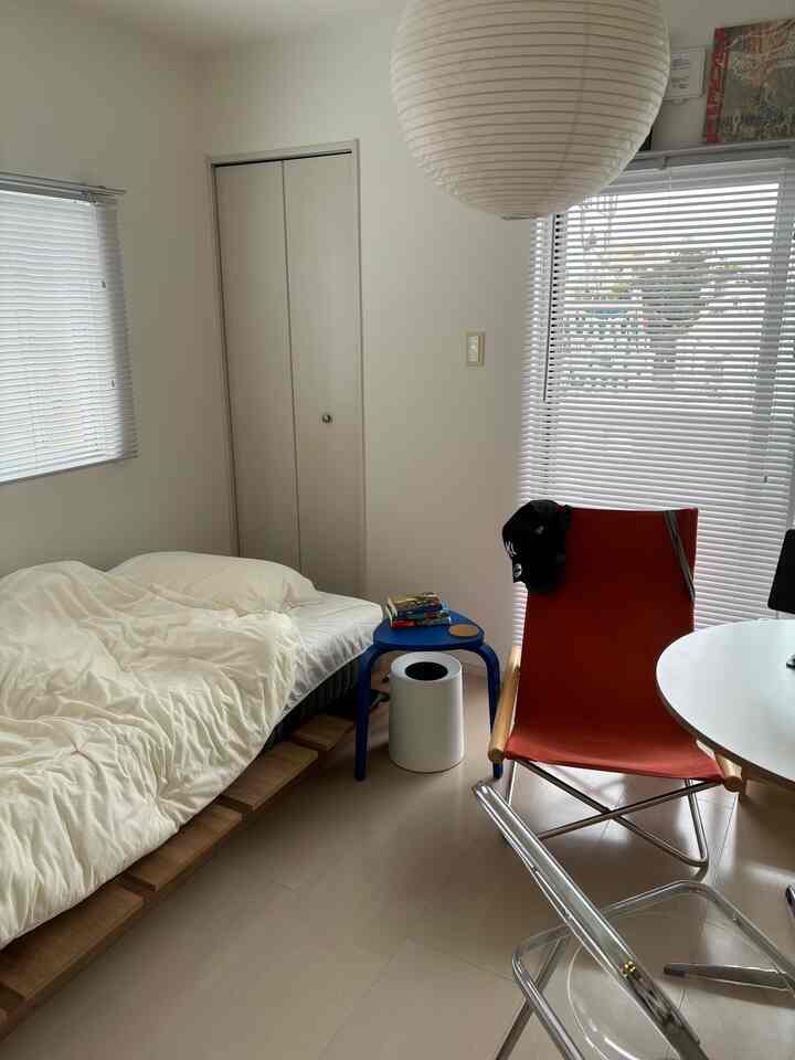 Bright white and wood-toned single-room bedroom featuring simple furniture, a blue stool, and an orange canvas armchair in a cozy setting