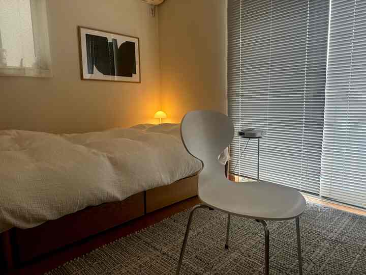 Natural wood-toned bedroom with warm lighting and blinds, featuring a white chair in a simple single household space