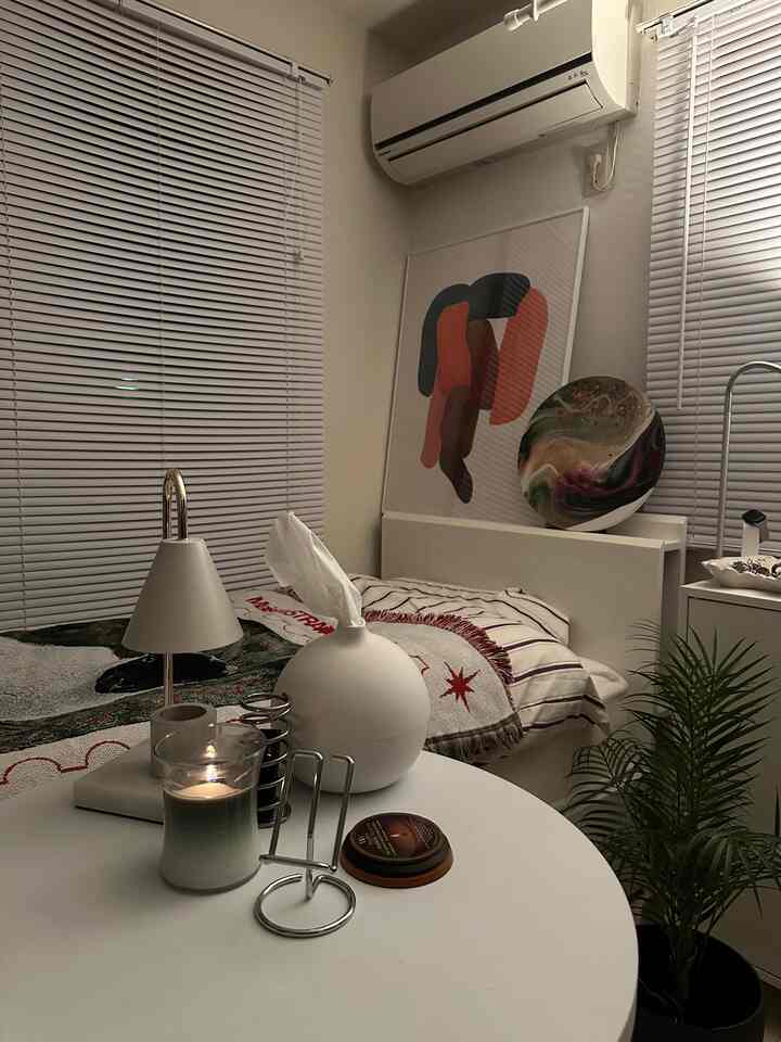 White and beige toned small bedroom featuring a single bed, lit candle on table, abstract poster above bed, and a potted plant, creating cozy atmosphere