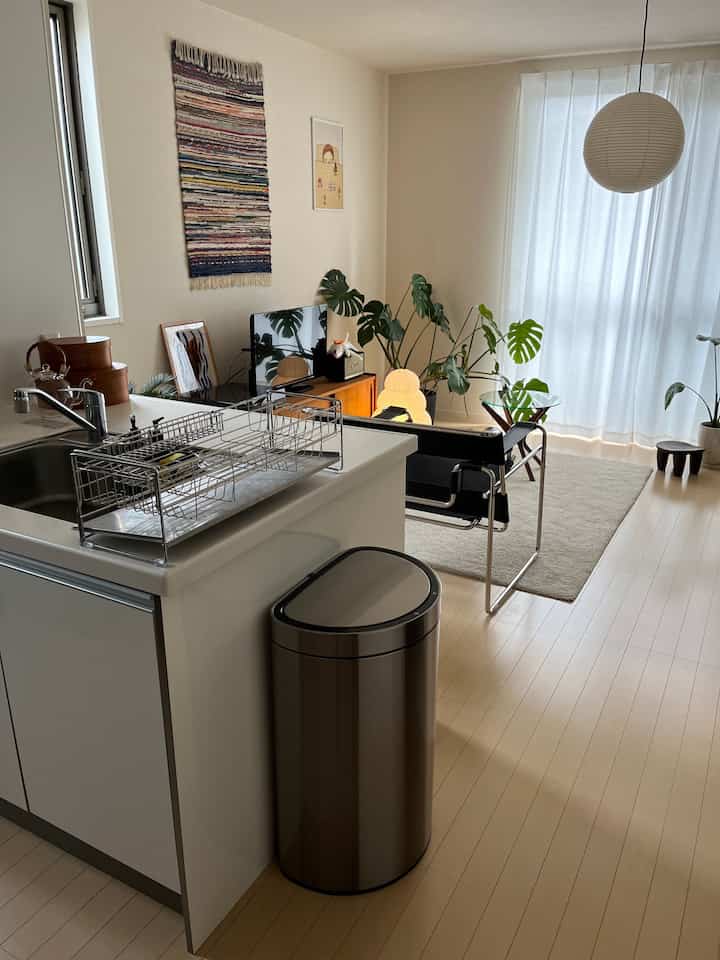 Natural color tones and white walls in a kitchen and living room area featuring a silver trash bin, black armchair, and pendant light in a clean interior