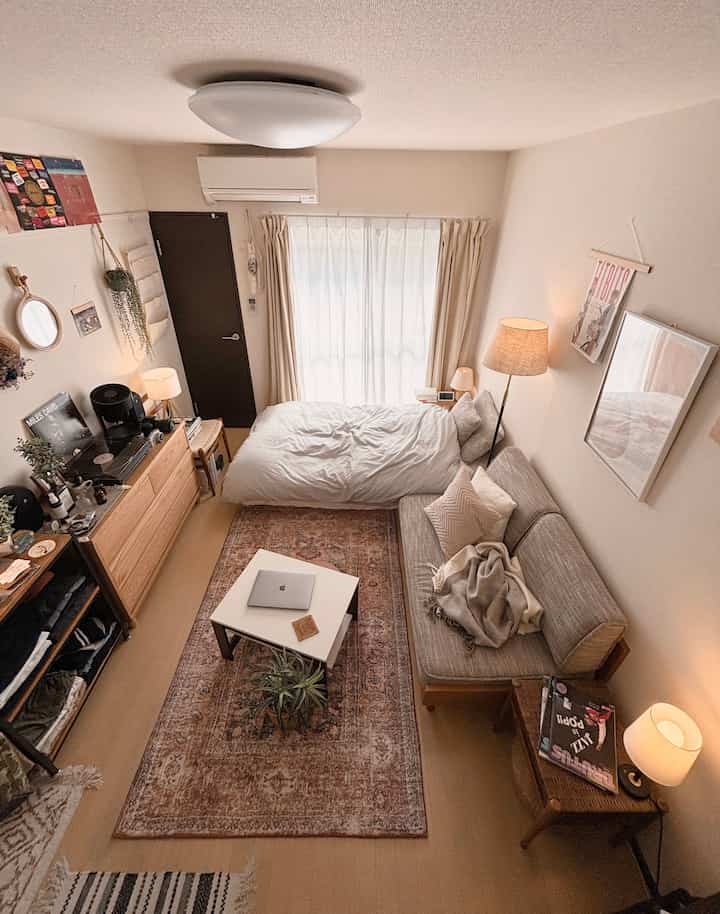 A beige natural-tone 1K studio living room featuring a bed, sofa, vintage rug, and indirect lighting creating a cozy atmosphere
