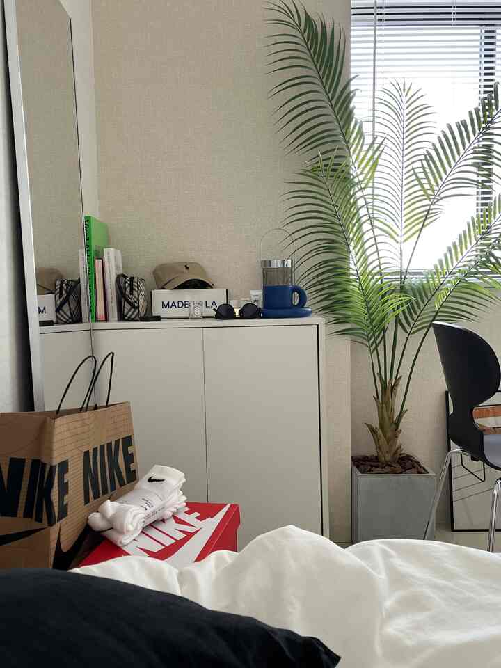 ベージュの壁とホワイトの家具が配置されたミニマルな寝室、グレーの鉢植えの大きな植物とブラックチェアがアクセントのあるインテリア