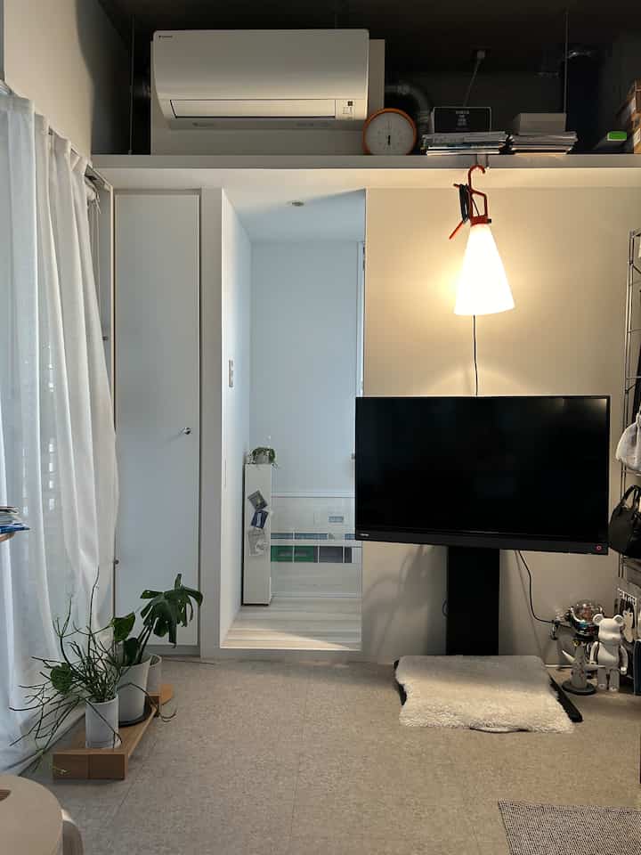 白を基調としたミニマルなワンルームのリビング、テレビスタンドと明るいフロアランプが配置された印象的な空間