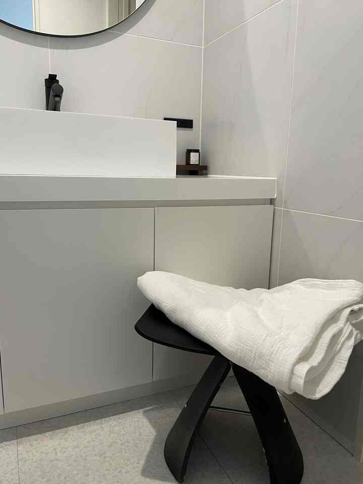 白と黒のトーンのバスルーム、洗面台とラウンドミラーが配置され、シンプルな黒いスツールと白いタオルが置かれている