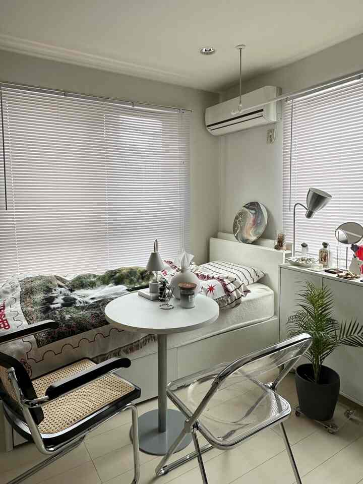 白を基調としたワンルーム、透明な椅子とラタンチェアのある丸いテーブルと清潔感のあるベッドが配置されたインテリア