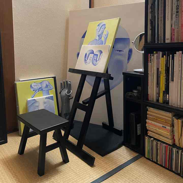 Natural tone small Japanese-style room featuring a black stool, easel with multiple paintings, creating a quiet artist workspace