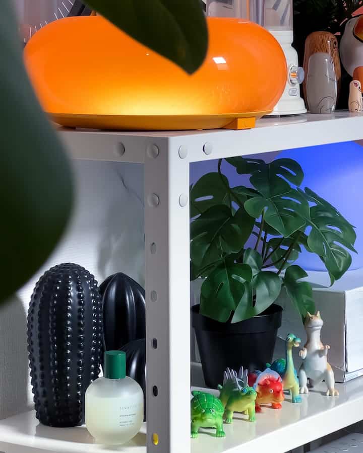 ホワイトメタルの棚に黒い植木鉢や多彩なオブジェ、オレンジのLEDライトが配置されたモダンでかわいらしいワークスペース