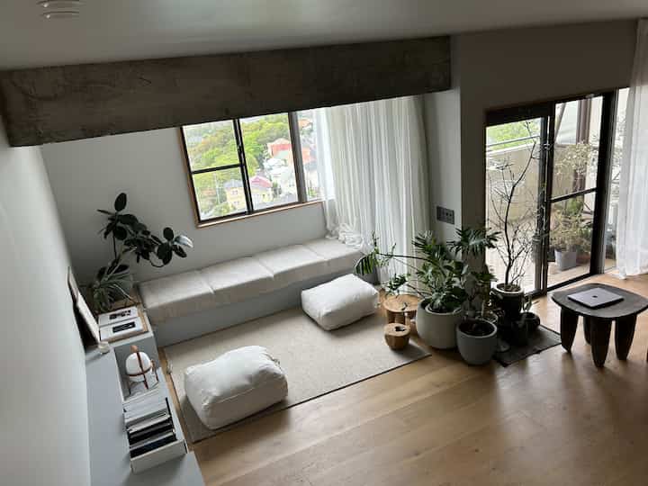 A white and wood tone living room by the window side featuring leafy plants and stools creating a cozy, natural atmosphere