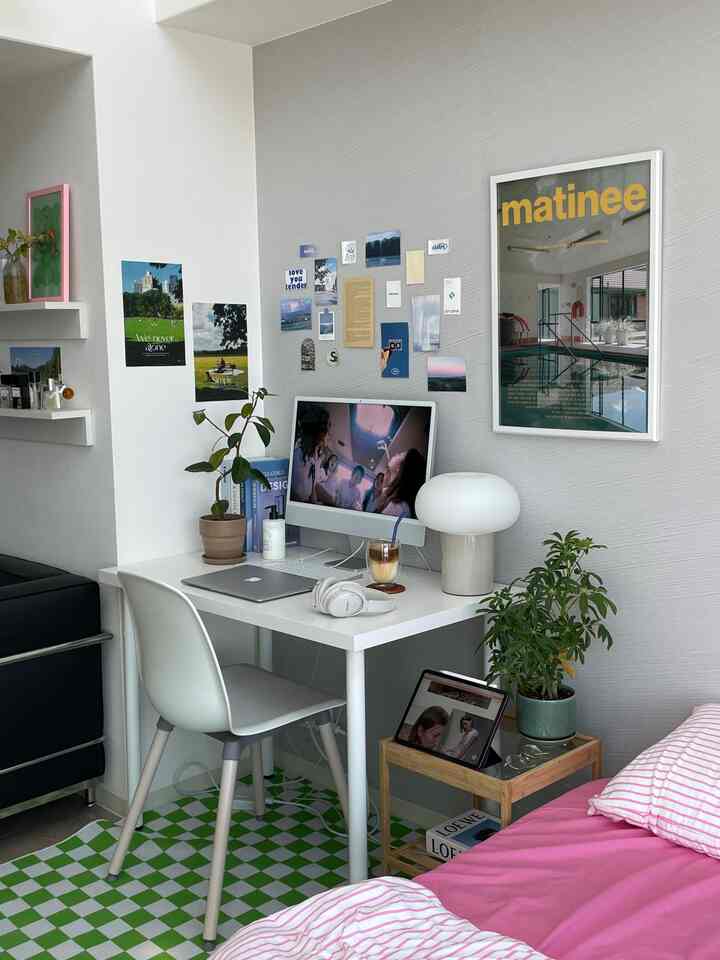 白を基調とした寝室兼ホームオフィス、白いデスクと椅子、ピンクの寝具、ポスター、植物が配された印象的な空間