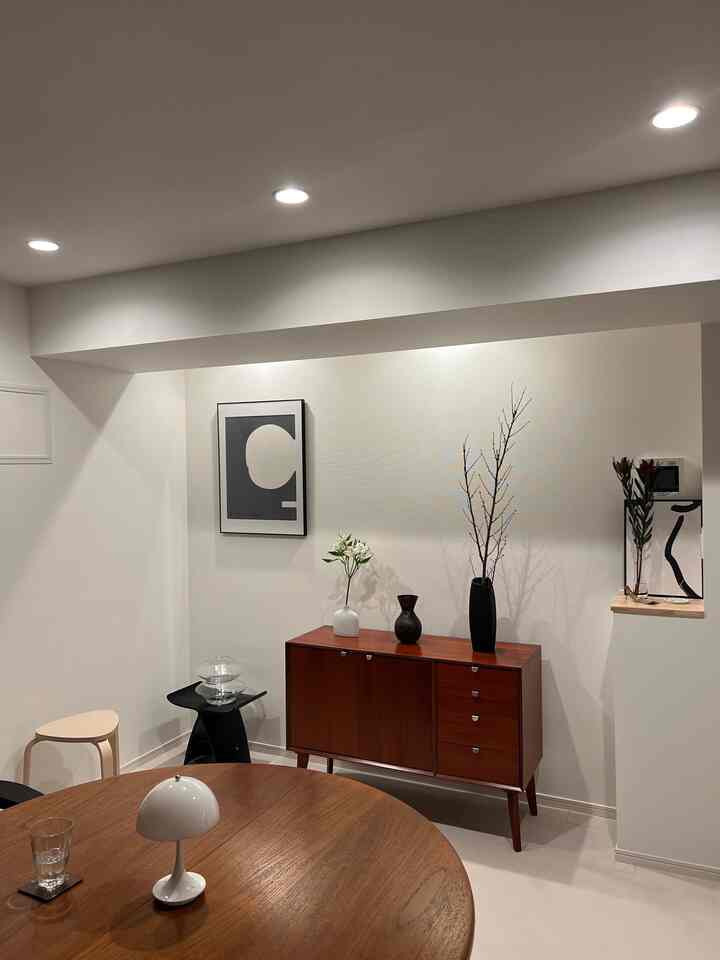 白い壁と天井のダイニングルーム、木製の丸いダイニングテーブルとミッドセンチュリーモダンのコンソールテーブル、テーブルランプと花瓶が配置されたシンプルなインテリア