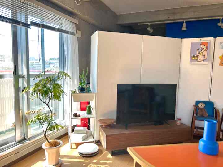 明るい白い壁と温かみのあるブラウンの木目調が調和したリビング、植物とテレビ台が配置された自然な雰囲気のインテリア