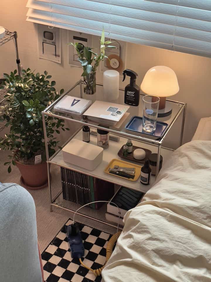 White and natural toned bedroom featuring a multi-tier nightstand with table lamp, diffuser, and organized trays creating a cozy bedside area