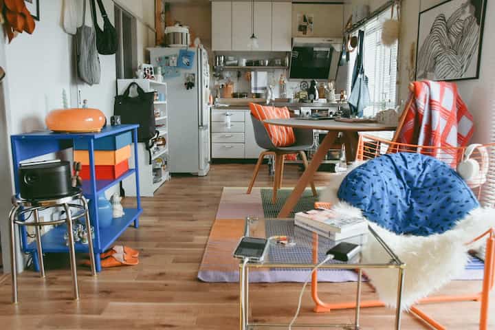 ナチュラルな色調の小さなワンルームキッチンとダイニングスペース、オレンジとブルーのアクセント家具が配置された雰囲気のあるインテリア