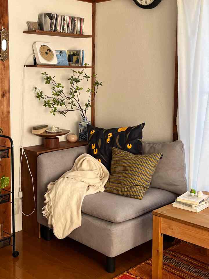 木目調とベージュトーンのリビングコーナー、グレーのアームチェアとクッション、ブランケット、植物が配置された落ち着いた雰囲気のインテリア