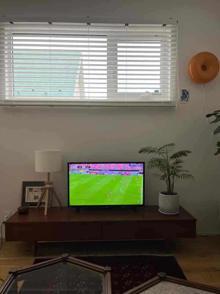 Bright white walls with a brown wood tone TV stand in a living room, complemented by green plant and table lamp creating a neat atmosphere