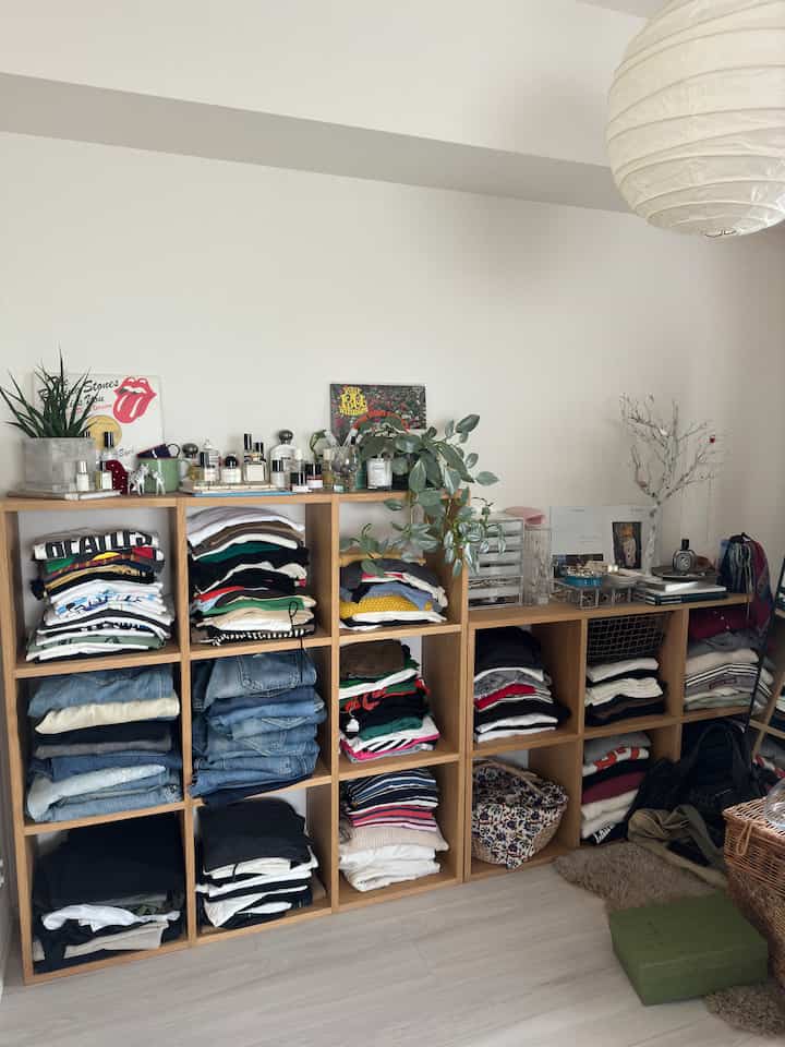 ナチュラルな色調の小さな部屋に、折りたたんだ服が整頓された木製の棚と観葉植物が配置された印象的な空間