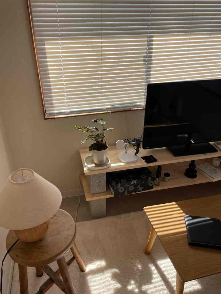 ベージュと木目調のナチュラルなリビング、ブラインド付き窓、テーブルランプ、テレビ台、ラグが配置された雰囲気のある空間