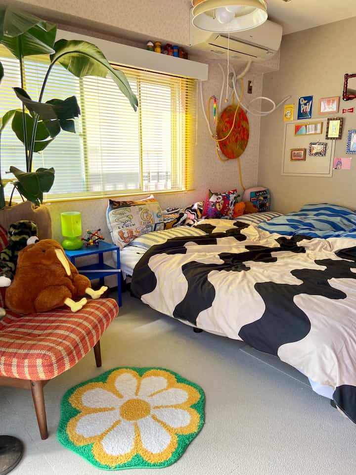 A small Showa Retro bedroom with bright yellow lighting and a green patterned rug, featuring uniquely patterned bedding and colorful cushions