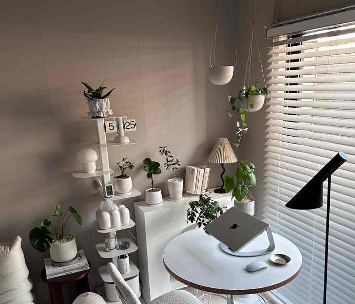 Bright white and green toned home office space featuring multiple plants on shelves and a round table with a laptop, creating a cozy atmosphere
