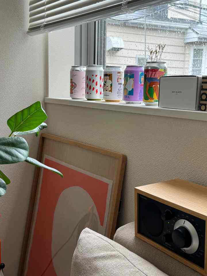 ナチュラルカラーの1LDKリビング、窓辺に多彩なデザインのビール缶が並び、ベージュのソファと木製ラジオが配置された雰囲気のあるインテリア