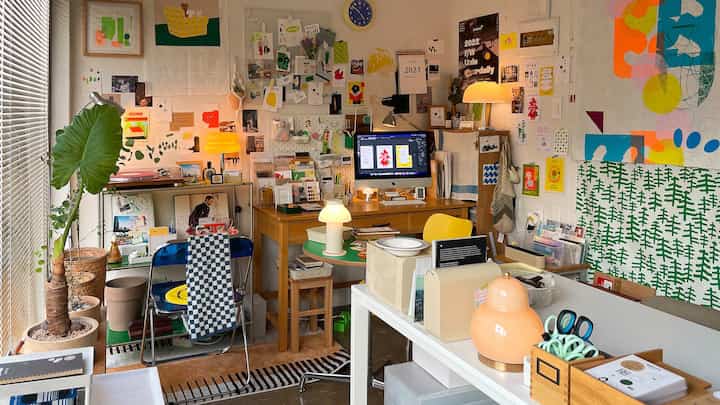 A vibrant home office featuring yellow and navy accents, multiple desks, plants, and an eclectic, cluttered yet creative atmosphere