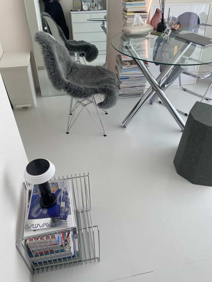 Simple modern dining room with white floor and glass dining table, featuring gray faux fur chairs and stacks of books