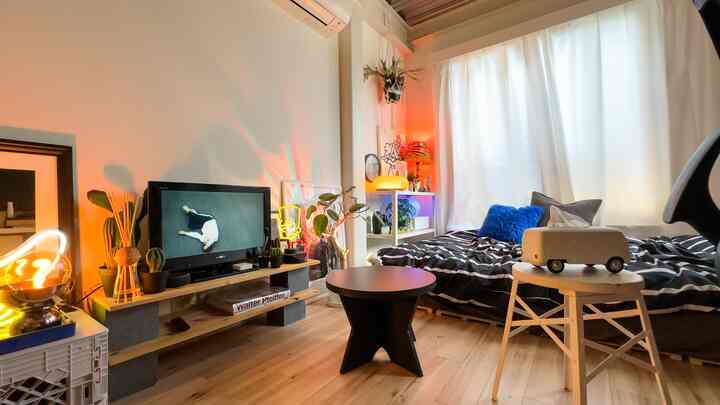 ナチュラルトーンの6畳ワンルーム、ベッドとテーブルランプ、テレビ台が配置された居心地の良い一人暮らし空間