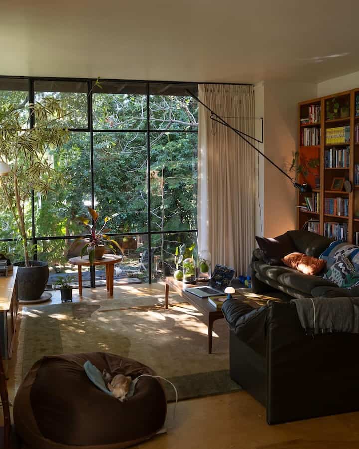 Natural-toned living room with wooden furniture, large glass windows offering forest views and plants creating a cozy atmosphere